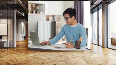 Side view of an Asian man working at a home desk using a tablet and taking notes. Wall mural