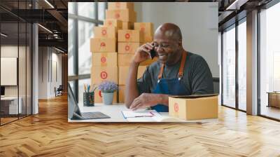 Smiling man in apron talking on phone, surrounded by boxes, working at desk with laptop and notebook. Wall mural