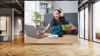 Smiling young asian freelancer woman is enjoying a healthy salad while working remotely on her laptop, embracing a balanced work from home lifestyle Wall mural