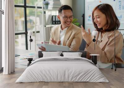 Two business partners smiling and discussing work while working together on a laptop in a modern office Wall mural