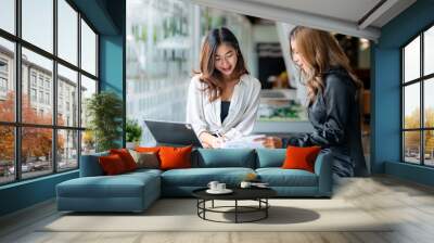 two pretty young asian businesswoman sitting at desk with laptop doing paperwork together discussing Wall mural