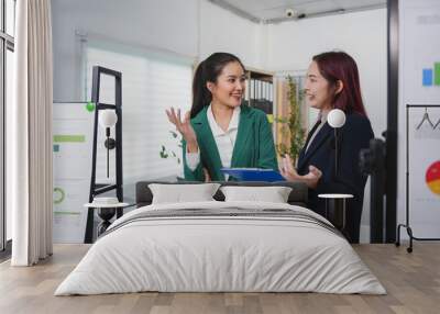 Two smiling businesswomen are discussing work in a modern office, using a clipboard and charts to analyze data and collaborate on a project Wall mural