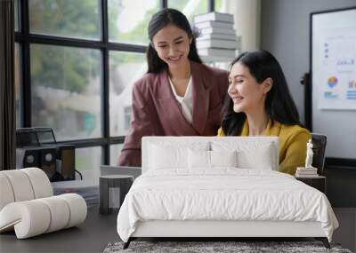 Two successful businesswomen collaborating joyfully on a laptop in a contemporary office, bathed in natural light Wall mural