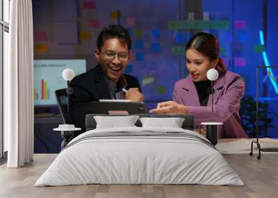 Two young business people smiling and discussing work strategy using digital tablet in modern office at night Wall mural