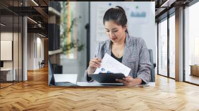 Young asian businesswoman working on paperwork and analyzing financial chart sitting at her desk in modern office Wall mural