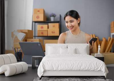 Young entrepreneur managing e-commerce orders from laptop and phone, surrounded by shipping boxes in home office setting. Wall mural