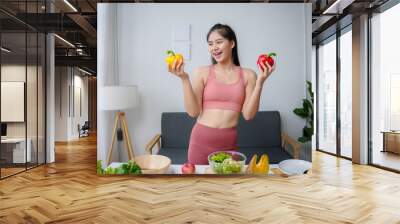 Young fitness woman is smiling while holding fresh bell peppers in a kitchen full of healthy ingredients Wall mural