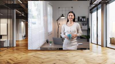 Young professional woman standing in modern home office holding a coffee cup, smiling confidently with a neat workspace in the background. Wall mural