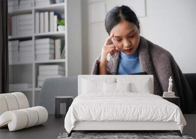 Young woman wrapped in a blanket, feeling sick and browsing the internet on her smartphone while sitting on a sofa Wall mural