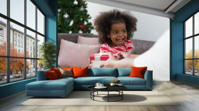 Cheerful cute African American little smiling girl receiving Christmas gift box or presents gift from adult hands at home. Christmas holiday celebration, Merry Christmas and Happy Holidays Wall mural