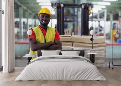 Confident smiling African American male warehouse worker in safety vest and helmet standing with arms crossed in automotive spare parts storage warehouse over parcel stack on forklift truck Wall mural