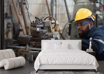 Male factory worker working at work maintenance machine in industrial factory while wearing safety uniform, glasses and hard hat. Black male technician and heavy steel lathe machine in workshop Wall mural