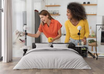 Two young woman cooking salad and playing with dog on table in kitchen room at home Wall mural