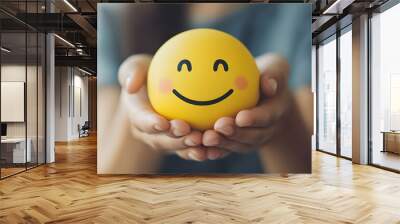 A woman holds a happy yellow smiley face emoji, symbolizing happiness, positive thinking, and a growth mindset. Perfect for celebrating World Smile Day, World Emoji Day and World Mental Health Day. Wall mural