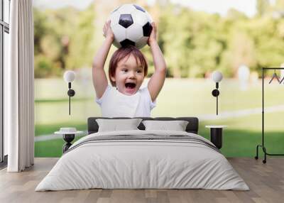 Excited little toddler boy playing football on soccer field outdoors: the kid is holding ball above head and shouting ready to throw it. Active childhood and sports passion concept Wall mural