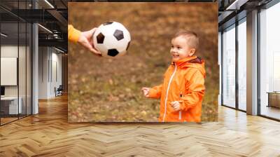 Happy little toddler kid eagerly watching at soccer ball in hand of father. Hand of dad giving to little son football ball in autumn forest or park. Family active leisure outdoors concept Wall mural