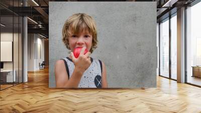 Adorable caucasian blond little 6 years old boy biting happy a red apple with vitamins looking to the camera outside isolated concrete background. Healthy eating in childhood concept. Copy space Wall mural