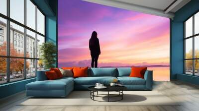 A woman stands on the beach at sunset, looking out at the ocean. The sky is a beautiful mix of pink and purple hues, creating a serene and peaceful atmosphere. The woman is lost in thought Wall mural