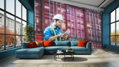 Man wears hardhat and reflection shirt and checking tablet with blurry metal containers in background. Concept of inventory and logistic management. Wall mural