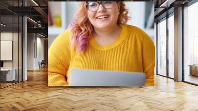 Modern electronic gadgets, job and occupation concept. Cute elegant young brunette female with excess weight using laptop computer for remote work, looking at screen, holding pen, writing down Wall mural