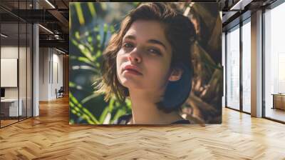 A woman with short, brown hair looks at the camera, surrounded by green leaves Wall mural