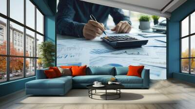 Person analyzing financial data with calculator and charts on desk for budgeting and accounting purposes in an office setting. Wall mural