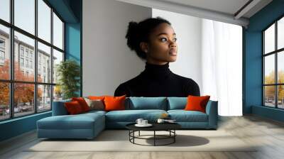 A contemplative black woman gazes out the window, showcasing a minimalist aesthetic with soft natural light highlighting her profile and serene expression Wall mural