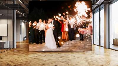 A crowd of young happy people with sparklers in their hands during celebration. Sparkler in hands on a wedding - bride, groom and guests holding lights in hand. Sparkling lights of bengal fires. Wall mural