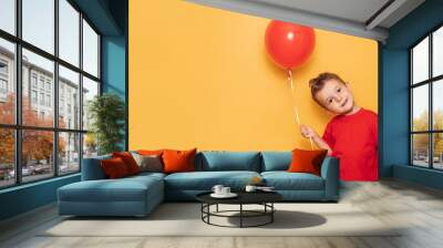 A happy Caucasian boy isolated on a bright yellow background holds a red balloon in his hands. A place for your text or advertisement. Wall mural