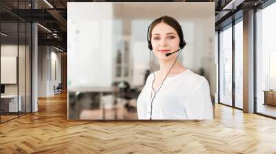 Friendly young female technical support dispatcher with a headset working in a call center on a hotline, talking on the phone. Portrait of an attractive customer care representative. Business concept Wall mural