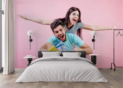 handsome young couple with plane tickets, boyfriend holding girlfriend imitating airplane isolated over pink Wall mural