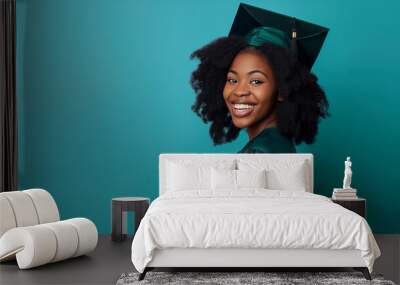 portrait of a happy young dark-skinned woman wearing a graduation gown and hat standing on a blue background, copyspace banner Wall mural