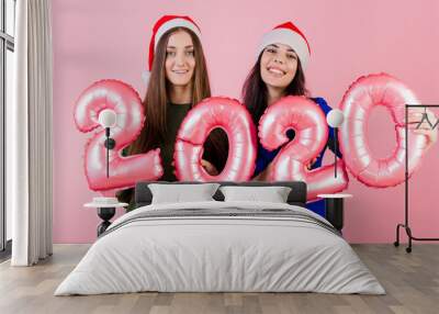 two smiling women wearing santa hats holding 2020 balloons for new year holiday isolated over pink Wall mural