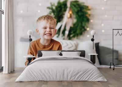 Cute happy Caucasian boy opening his presents on Christmas morning. Christmas tree wreath on background. Child excited and smiling. Happy family holidays at home. Opening gifts in boxes. Copy space. Wall mural