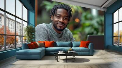 freelancer work concept.A Black man with medium-length hair and a beard, wearing a grey sweater, is working on his laptop in an urban setting with plants around him.  Wall mural