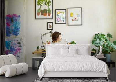 Female artist at her workplace working from home. Young woman dressed in jeans and striped shirt sitting at the table and creating an illustration. Wall mural