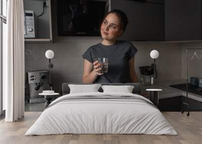 A young brunette woman stands in the kitchen and drinks water from a glass glass Wall mural