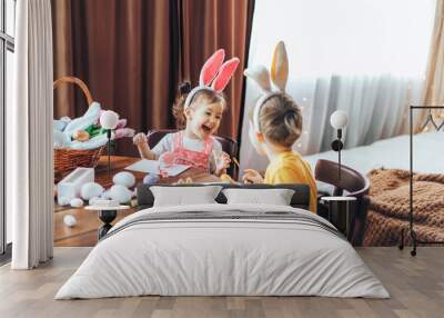 A boy and a girl are painting Easter eggs for the holiday at a wooden table. Children have bunny ears on their heads Wall mural