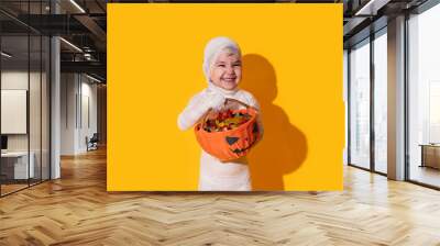 Child in mummy costume holding basket of chocolates in front of yellow background Halloween trick or treat Wall mural