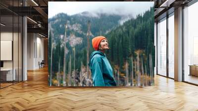 Lady with an orange hat stands in front of mountains with green fir trees. Wall mural