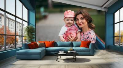 Young beautiful mother with a small child sitting on a bench in a park in pink clothes Wall mural