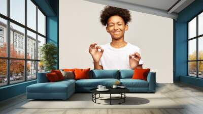 African american teen kid wearing white mockup t-shirt with copy space doing yoga, keeping eyes closed, holding fingers in mudra gesture. Afro boy showing ok sign with two hands on gray background Wall mural