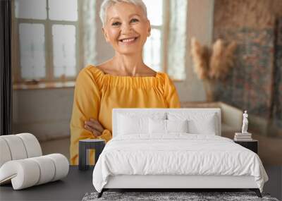 Attractive confident mature blonde woman wearing yellow dress crossing arms on chest and smiling happily at camera, posing in her newly renovated apartment with sofa and windows in background Wall mural