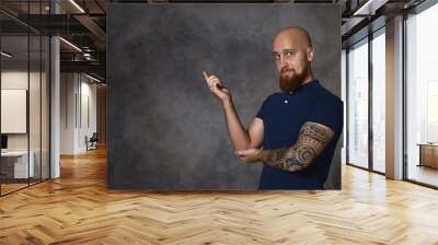 Attractive stylish European male with bushy beard, bald head and tattoo posing isolated in studio, pointing index finger, indicating copy space on gray wall for your advertisement or information Wall mural