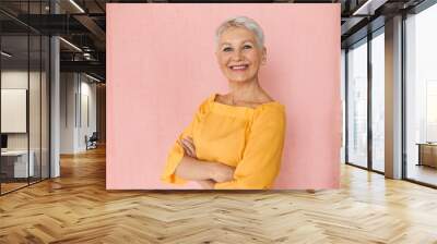 Attractive successful middle aged blonde businesswoman with short pixie hair and charming confident smile posing isolated against blank pink wall background, keeping arms crossed on her chest Wall mural