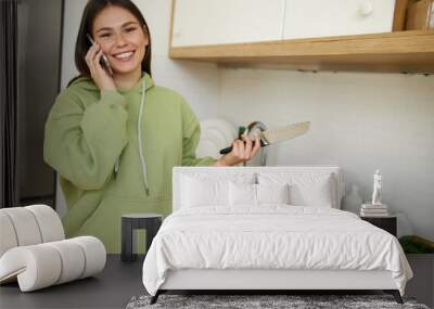 Beautiful positive young woman holding knife, slicing organic vegetables for salad, making lunch while talking on smart phone, smiling at camera. Cooking, housework and electronic gadgets concept Wall mural