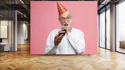 Celebration and anniversary concept. Emotional senior man with thick gray beard dressed in fancy elegant clothes, holding birthday cake and touching face in shock, stunned with surprise party Wall mural