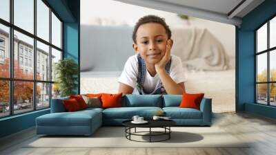 Childhood, lifestyle and positive human expressions. Cute charming dark skinned African schoolboy lying on his stomach on carpet in cozy bedroom holding hand under his chin, smiling joyfully Wall mural