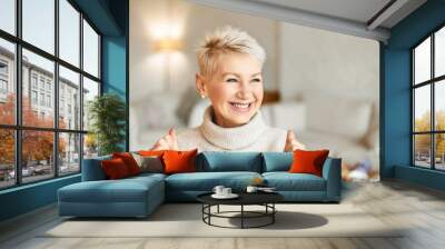 Close up portrait of beautiful middle aged woman with short blonde hair and radiant smile rejoicing at good news, being in festive mood, keeping her fists clenched, expressing joy and excitement Wall mural