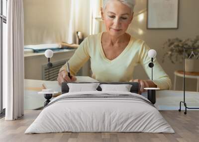 Concentrated middle aged woman with short hair calculating income and expenses, paying domestic bills, writing down in journal, sitting at table. People, finance, economy and paperwork concept Wall mural